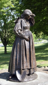 Bronze statue of pregnant female historical figure Elizabeth Thorn wiping her brow and carrying shovel at her side, with a green cemetery in the background.