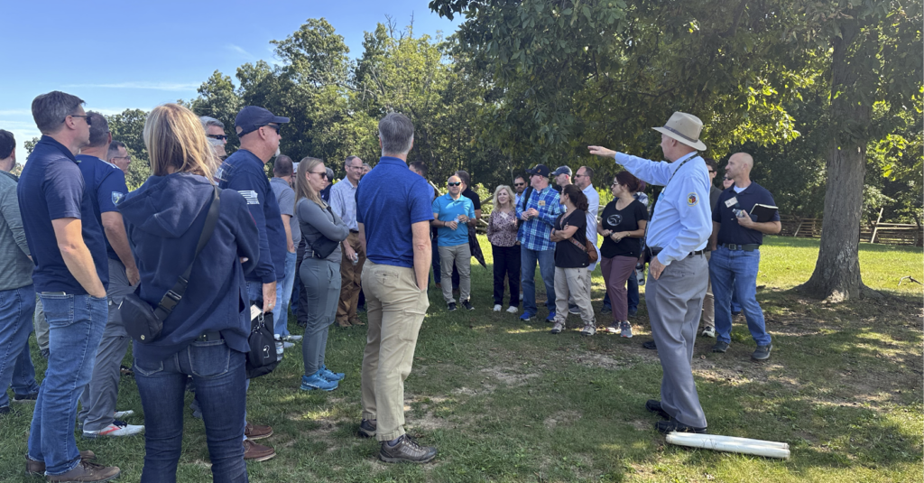 Group of people in a shaded, grassy setting listen to battlefield guide